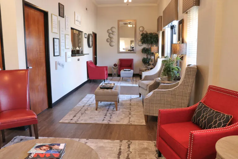 PCA NWA mental health services - A modern waiting room with red and patterned armchairs, a coffee table, wall art, plants, and large windows letting in natural light.