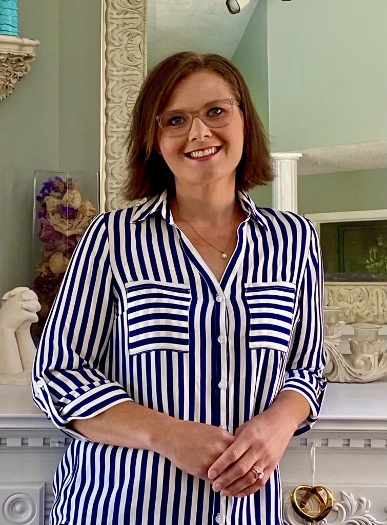 PCA NWA mental health services - Woman with short brown hair, glasses, and a blue and white striped shirt stands indoors in front of a white ornate fireplace mantel, smiling at the camera.