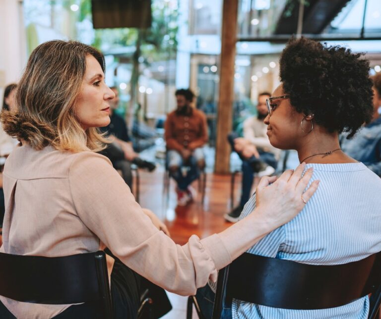 PCA NWA mental health services - Two women sit in a support group setting, with one offering comfort as the other discusses her experience with EMDR Therapy. Other people are visible sitting in a circle in the background.