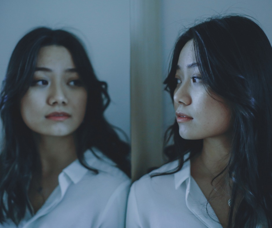PCA NWA mental health services - A woman with long dark hair and a white shirt looks at her reflection in a mirror, both faces partially illuminated, evoking the introspection often experienced during EMDR Therapy.