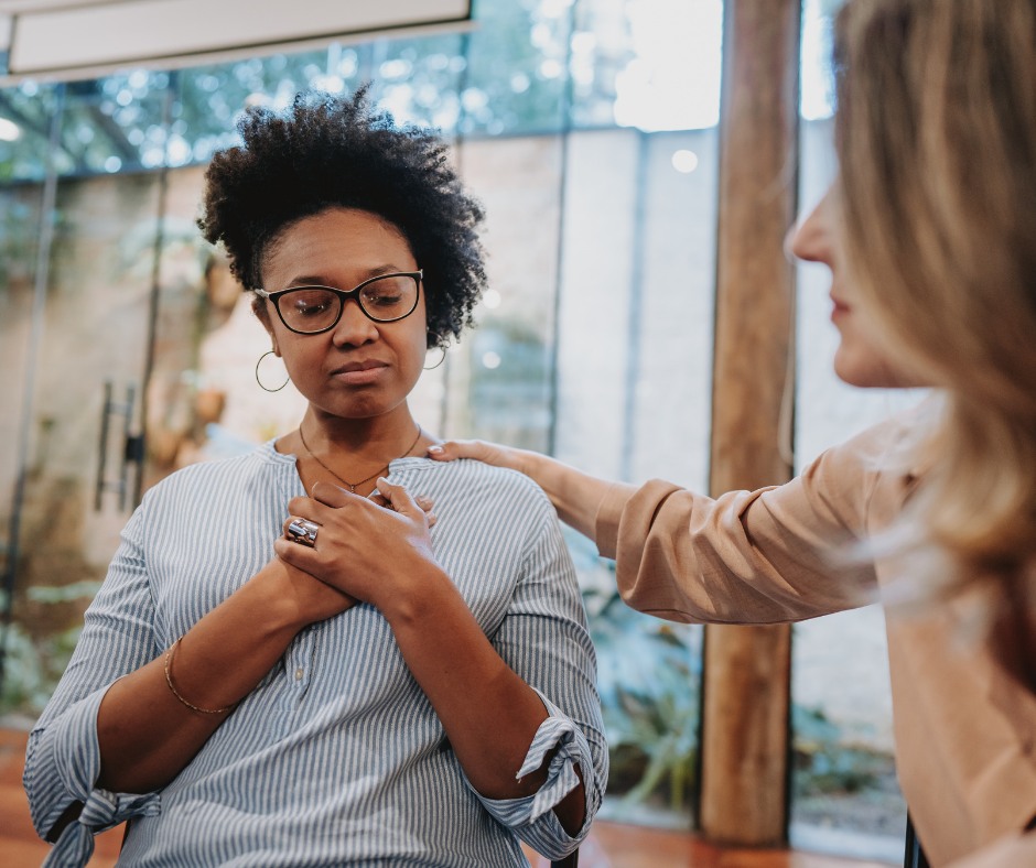 PCA NWA mental health services - Two women sit indoors; one looks emotional with her hands on her chest while the other places a comforting hand on her shoulder, illustrating the supportive environment of EMDR Therapy.