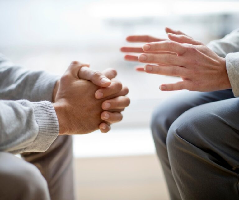 PCA NWA mental health services - Two people sit facing each other; one has hands clasped while the other gestures with open hands, suggesting a conversation or discussion, possibly within an EMDR Therapy session.