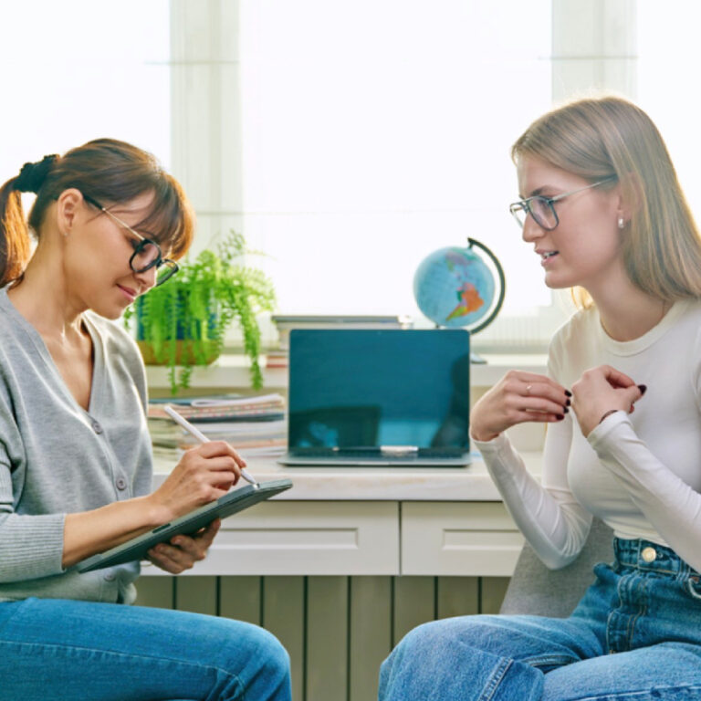 PCA NWA mental health services - Two women sit facing each other in an office. One takes notes on a clipboard, while the other gestures as she discusses EMDR Therapy. A laptop, globe, and plant are on a desk in the background.