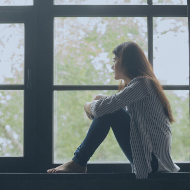 PCA NWA mental health services - A woman sits barefoot on a windowsill with her knees up, gazing outside through a large window. She appears lost in thought, as if reflecting after an EMDR Therapy session. Lush trees are visible in the background.