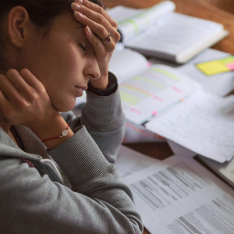 PCA NWA mental health services - A person sits at a desk with their hand on their forehead, eyes closed, surrounded by open books and papers, appearing stressed or tired while studying—feelings that EMDR Therapy may help address.