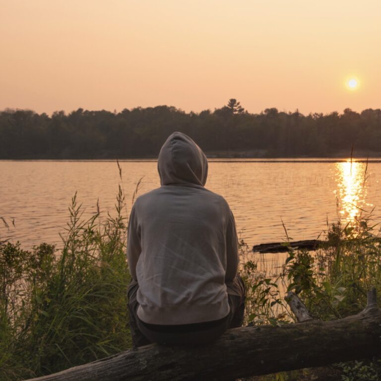 PCA NWA mental health services - A person in a hoodie sits on a log facing a lake at sunset, with trees and plants in the foreground and background.