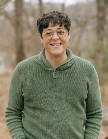 PCA NWA mental health services - A social worker with short dark hair and glasses, wearing a green sweater, stands outdoors in a wooded area, smiling at the camera.