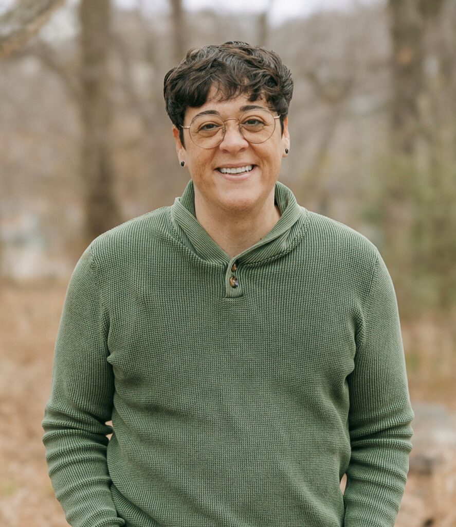 PCA NWA mental health services - A social worker with short dark hair and glasses, wearing a green sweater, stands outdoors in a wooded area, smiling at the camera.