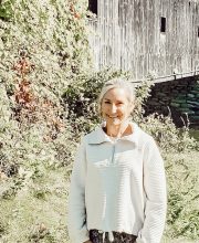 PCA NWA mental health services - A woman in a light sweater stands outdoors in front of a wooden building surrounded by greenery, evoking the tranquil setting often encouraged in adult psychiatry for relaxation and mental well-being.