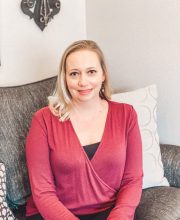 PCA NWA mental health services - A woman with blonde hair wearing a red long-sleeve top sits on a gray armchair with patterned cushions, facing the camera.