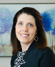 PCA NWA mental health services - Woman with shoulder-length brown hair wearing a black blazer and patterned top, smiling in front of a blue and purple abstract painting.