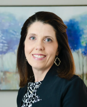 PCA NWA mental health services - Woman with shoulder-length brown hair wearing a black blazer and patterned top, smiling in front of a blue and purple abstract painting.