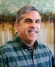 PCA NWA mental health services - A middle-aged man with short gray hair wears a green plaid shirt and smiles at the camera, standing in front of a blurred nature-themed background.