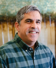 PCA NWA mental health services - A middle-aged man with short gray hair wears a green plaid shirt and smiles at the camera, standing in front of a blurred nature-themed background.