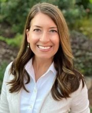 PCA NWA mental health services - A woman with long brown hair wearing a white shirt and light cardigan smiles outdoors with greenery in the background.
