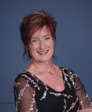 PCA NWA mental health services - A woman with short reddish-brown hair smiles, standing with arms crossed, wearing a black dress with floral patterns, in front of a blue background.