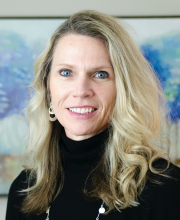 PCA NWA mental health services - A woman with long blonde hair and blue eyes wearing a black turtleneck and silver earrings stands in front of a colorful, blurred painting.