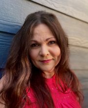 PCA NWA mental health services - Woman with long brown hair wearing a sleeveless red top stands in front of a blue wooden wall, looking at the camera with a neutral expression.