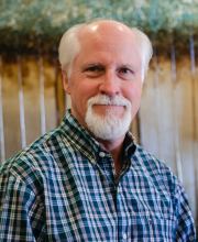 PCA NWA mental health services - An older man with a white beard and mustache, wearing a green plaid shirt, stands indoors with a blurred background.