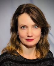 PCA NWA mental health services - A woman with shoulder-length brown hair and blue eyes, wearing a black striped top, poses against a plain light background.