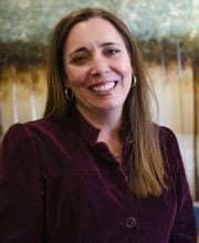 PCA NWA mental health services - A woman with long brown hair, wearing a maroon jacket and hoop earrings, smiles at the camera in an indoor setting with a blurred background.