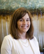 PCA NWA mental health services - A woman with shoulder-length brown hair, wearing a white top and pearl necklace, smiles at the camera. A blurred, abstract background is visible behind her.