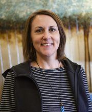 PCA NWA mental health services - A woman with brown hair, wearing a black vest over a striped shirt and a turquoise pendant necklace, stands in front of an abstract background.