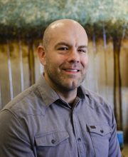 PCA NWA mental health services - A man with a shaved head and trimmed beard, wearing a gray button-up shirt, smiles at the camera with a blurred natural background behind him.