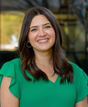 PCA NWA mental health services - Alyssa Raggio, Psy.D. smiles while standing outdoors in front of a glass building, radiating confidence and positivity—an image that beautifully captures the essence of psychology in everyday life.