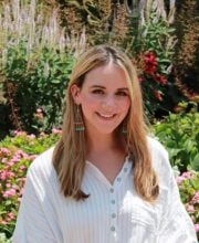 PCA NWA mental health services - A woman with long blonde hair, wearing a white shirt and colorful earrings, stands outdoors in front of a garden with pink flowers and greenery.