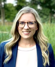 PCA NWA mental health services - A woman with long blonde hair and glasses, wearing a blue blazer and white top, stands outdoors in front of greenery, smiling at the camera.