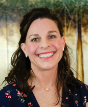 PCA NWA mental health services - A woman with wavy brown hair and a navy floral top smiles at the camera, standing in front of a blurred outdoor-themed background.