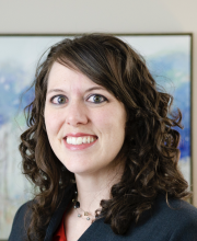 PCA NWA mental health services - Woman with curly brown hair wearing a dark blazer and necklace, smiling at the camera with a blurred, colorful painting in the background.