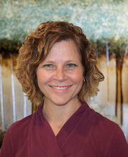 PCA NWA mental health services - A woman with short curly hair wearing a maroon top is smiling in front of a blurred outdoor background with green and brown tones.
