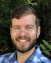 PCA NWA mental health services - A man with short brown hair and a beard, wearing a blue plaid shirt, smiles outdoors with green foliage in the background.