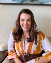 PCA NWA mental health services - Woman with long brown hair sits and smiles, wearing a white shirt with orange and yellow patterns and a brown belt, with a candle visible on a table beside her.